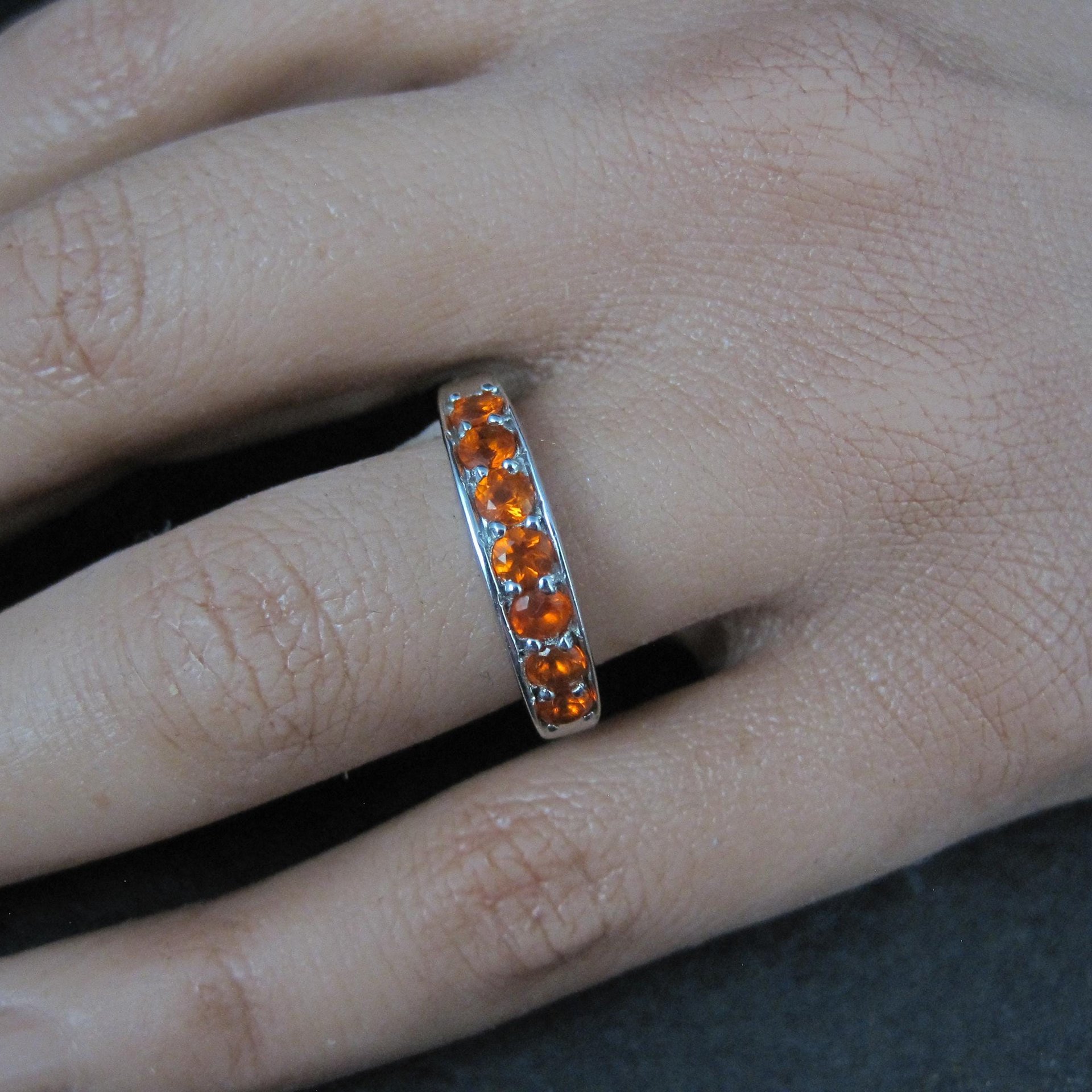 May include: A silver ring with a band of orange gemstones. The ring is worn on a finger, showcasing the bright, eye-catching color of the stones. The gemstones are evenly spaced around the ring.