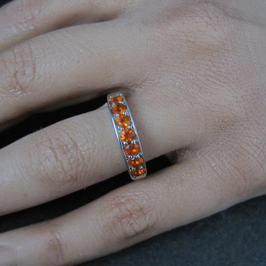 May include: A silver ring with a band of orange gemstones. The ring is worn on a finger, showcasing the bright, eye-catching color of the stones. The gemstones are evenly spaced around the ring.